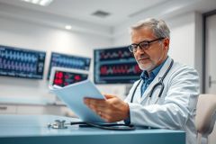Mature doctor in a bright, modern hospital reviewing patient charts. He wears glasses and a white coat, with medical equipment and monitors visible in the background. Healthcare, medical professional, patient care, diagnostics.