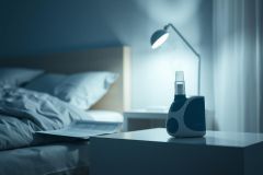 Blue-toned bedroom scene featuring a nebulizer machine on a nightstand illuminated by a lamp. The image depicts a restful atmosphere, possibly related to respiratory health, sleep, or medical devices. Soft lighting and a blurred bed create a calming mood.