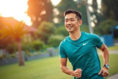 The image shows a man wearing a sports outfit, including a dark teal or aqua-colored shirt and shorts, running along an asphalt path. He is smiling and appears to be enjoying the activity.