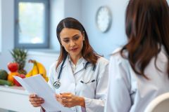 A female doctor in a white coat examines paperwork while a patient sits opposite. The bright, modern office features fruit and a window. Healthcare, consultation, patient care.
