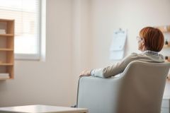 A woman sits in a gray armchair in a bright, minimalist therapy office. The scene depicts mental health, counseling, or a consultation. Warm natural light from a window illuminates the room with neutral tones and a wooden bookshelf in the background. Focus on introspection and well-being.