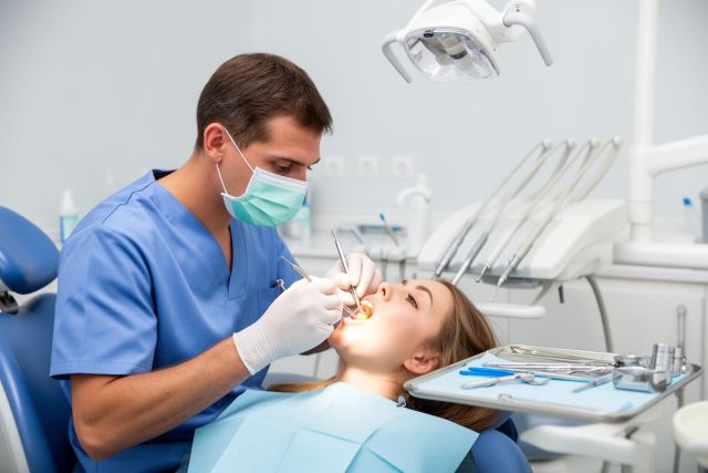 Dentist in scrubs and mask examining a reclined patient's mouth with a dental mirror and probe.
