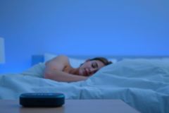 Woman sleeping peacefully in a blue-toned bedroom. A smart speaker sits on a nightstand beside the bed, suggesting sleep sounds or relaxation aids. Focus on restful sleep and modern bedroom technology.