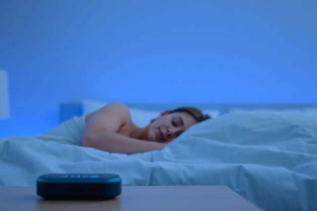 Woman sleeping peacefully in a blue-toned bedroom. A smart speaker sits on a nightstand beside the bed, suggesting sleep sounds or relaxation aids. Focus on restful sleep and modern bedroom technology.