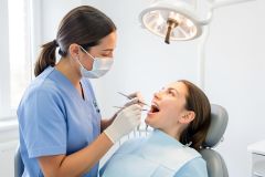 Dental hygienist wearing a mask examines a patient's open mouth under an exam light during a routine gum check.