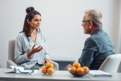 Doctor consults with a patient in a bright, modern office setting. The image features a female healthcare professional and an older man, suggesting a medical discussion. Colors are predominantly neutral with pops of orange from fruit bowls. Focuses on patient care and healthcare services.