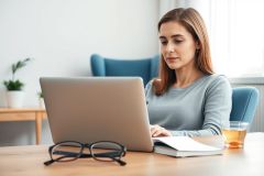 Woman working on laptop at home. Bright, modern living room setting with a wooden table, glasses, notebook, and plants. Focus on remote work, technology, and a comfortable home office environment. Cool blue and neutral tones.