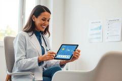 A smiling female doctor in a white coat uses a tablet in a bright, modern medical office. Charts are visible on the wall. Focus on healthcare technology, patient care, and a professional work environment. Blue and white color scheme.