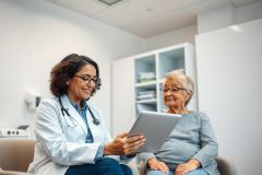 Doctor consults with elderly patient in a bright, modern clinic. The healthcare professional uses a tablet during the appointment, showcasing digital health and senior care. Warm lighting and neutral tones create a comforting atmosphere.
