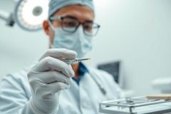Doctor in surgical scrub cap and mask holds a dental tool in a sterile, bright operating room. Focus on the instrument, suggesting dental procedures or medical treatment. Cool blue and white tones dominate the scene.