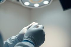 Close-up of gloved hands holding a dental implant in a bright, modern dental office. Focus on medical equipment and precision dentistry. Cool blue and white tones create a clinical, professional atmosphere.
