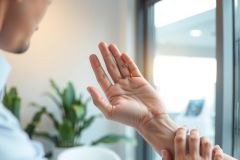 Close-up of a person holding their wrist, possibly experiencing pain or discomfort. Warm, natural lighting highlights the skin tone and textures. Focus on hand and wrist with blurred background.