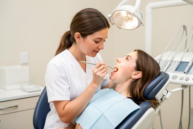 Dental clinician performs an oral exam with a mirror while a patient reclines in a dental chair.