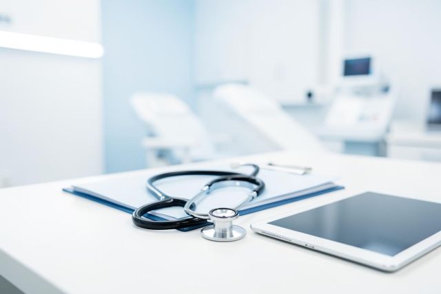 A professional medical stethoscope, digital tablet, and blue folder rest on a clean white desk in a bright, modern hospital or clinic setting. The blurred background features medical equipment in soft blue and white tones, emphasizing a sterile and high-tech healthcare environment.