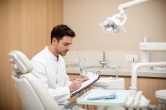 Dentist reviewing patient notes on a clipboard beside a dental chair in a clean clinic.