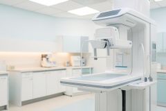 High-tech white medical imaging machine in a bright, modern radiology clinic. This diagnostic mammography equipment is set within a clean, sterile hospital environment featuring white cabinetry and soft blue tones. The professional healthcare setting emphasizes advanced medical technology and clinical precision.