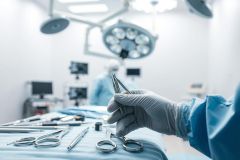 Surgical scene in a modern operating room. A gloved hand holds surgical instruments amidst a backdrop of sterile blue linens, medical equipment, and bright overhead lights. Focus on precision and medical procedures.
