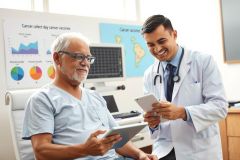 The image shows a medical professional, wearing a white lab coat, in conversation with an elderly patient. The setting appears to be a clinical or hospital environment, with a desk and medical equipment visible. Both individuals are seated; the doctor is standing next to the patient, who has a slight smile on his face while holding a clipboard. The scene suggests a friendly interaction during a medical consultation.