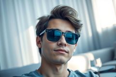 Stylish young man wearing black sunglasses indoors. Portrait with a blurred background featuring light gray curtains. Casual, modern look with a focus on eyewear and a relaxed atmosphere.