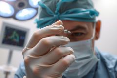 Close-up of a surgeon's gloved hand holding a surgical instrument in an operating room. Medical professional wearing a mask and surgical cap. Focus on precision and healthcare.