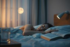 Woman sleeping peacefully in a blue-toned bedroom at night. An open book rests on the bed beside her, illuminated by a bedside lamp with moonlight filtering through curtains. Serene, restful, and cozy bedroom scene.