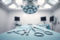 Surgical instruments laid out on a sterile blue operating table in a modern operating room. Bright white surgical lights illuminate the scene with monitors visible in the blurred background. Focus on medical tools and a clean, clinical environment. Healthcare, surgery, hospital theme.