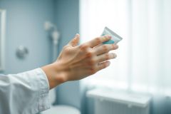 Close-up of a hand holding a tube of skincare cream in a bright, modern bathroom. Soft blue and white tones create a clean, clinical aesthetic. Focus on skincare, beauty products, hygiene, and wellness.