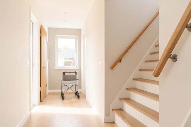 A bright and modern home hallway featuring a silver rollator walker positioned near a window. The space includes light wood stairs with handrails on the right and light wood flooring. The interior is decorated in neutral cream and white tones, filled with natural sunlight, emphasizing accessibility and mobility in a clean, minimalist living environment.