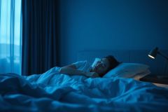 Woman sleeping peacefully in a blue-toned bedroom at night. Soft lighting from a bedside lamp illuminates the scene, creating a calm and restful atmosphere. Focus on sleep, relaxation, and nighttime tranquility.