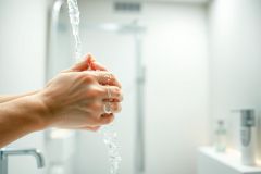 Close-up of hands washing under running water in a modern bathroom. Clean, bright, and emphasizing hygiene and sanitation. White and neutral tones with a focus on water droplets.