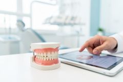 A professional dental model of teeth and gums rests on a white surface next to a digital tablet being operated by a hand. The background features a bright, blurred dental office with medical equipment. This scene illustrates dental consultation, oral hygiene education, and modern digital dentistry in a clean, clinical setting.
