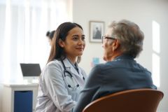Doctor consulting with a patient in a medical office. A female physician in a white coat listens attentively while the patient sits facing her. The scene shows a healthcare setting with a warm, professional atmosphere.
