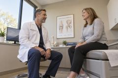Orthopedist consulting a middle-aged patient in exam room with joint anatomy poster behind.