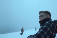 The image captures a moment in a foggy, snowy landscape. A young man, with light skin and short hair, is seated on the right side of the frame. 