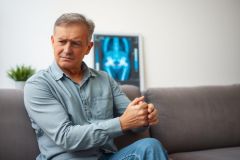 The image shows a man seated on a couch in a living room setting. The man is wearing a light gray shirt and jeans. He has his arm resting against the side of his body, possibly indicating discomfort or pain.