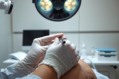Close-up of a medical professional wearing gloves performing a procedure, likely surgery, under bright operating room lights. Focus is on hands and equipment in a sterile environment.