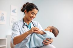 A smiling female doctor in a white coat and stethoscope examines a newborn baby wrapped in a blue blanket. The scene takes place in a bright, medical office setting with soft blue walls and pediatric artwork. Focuses on infant care and healthcare professionals.
