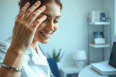 Smiling woman in a white coat, likely a doctor or healthcare professional, stands near a laptop in a modern office setting. Bright, clean environment with soft lighting. Focus on professionalism and medical context.