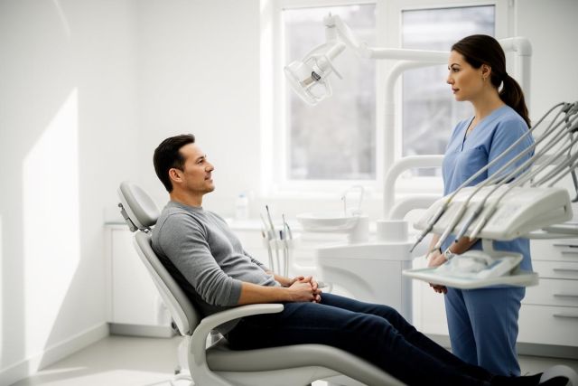Man reclines in a dental chair talking with a dental professional in a bright, clinical exam room.