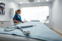 Medical examination room with surgical instruments on a blue exam table. A nurse in scrubs is blurred in the background. Clean, bright interior with a focus on healthcare and medical procedures. Blue and white color scheme, showcasing a clinical environment.