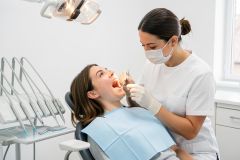 Dentist wearing mask and gloves examines a patient's open mouth in a bright, professional dental clinic.