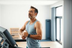 Man exercising on a treadmill in a modern home gym. He’s wearing a grey tank top and checking his fitness tracker. Bright, clean interior with white walls and large windows. Focus on fitness, healthy lifestyle, and indoor workout.
