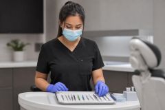 Dental clinician wearing mask and gloves arranging instruments on tray beside dental chair.