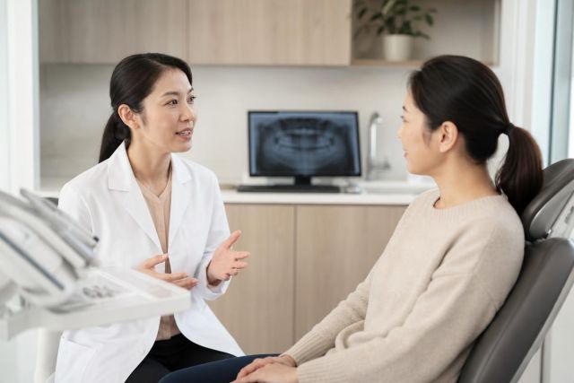 Dentist consulting seated patient in dental operatory with dental X-ray visible on monitor.