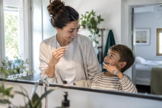 Mother and young son smiling while brushing teeth together at a bright bathroom sink.