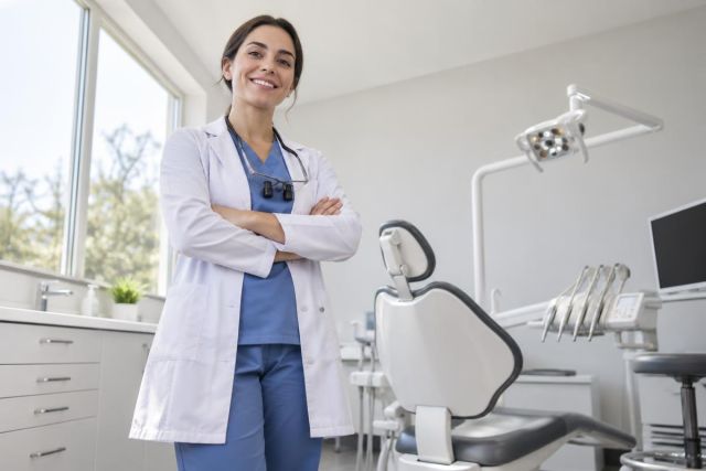 Smiling dentist in clinic standing beside dental chair and instruments, conveying trust and professionalism.