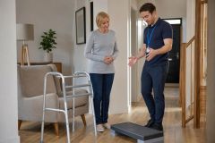 Home physiotherapist demonstrating step exercise to older woman preparing for hip surgery recovery.