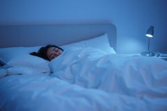 Woman sleeping peacefully in a cozy bedroom under soft blue lighting. A bedside lamp illuminates a white bed with fluffy pillows and blankets, creating a serene and restful atmosphere. Focus on sleep, relaxation, and nighttime ambiance.