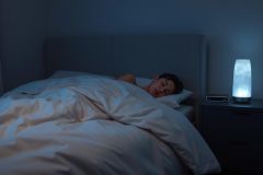 A young person sleeps peacefully in a dimly lit bedroom. A glowing, blue-toned lamp sits on a nightstand, creating a calming atmosphere. The image evokes sleep, relaxation, and a cozy bedroom setting. Blue and white tones dominate the scene.