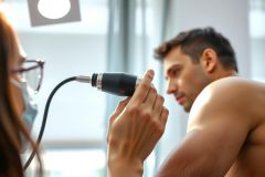 Doctor examining a patient's ear with an otoscope during a medical check-up. The patient is wearing a mask. Bright, well-lit clinic setting.